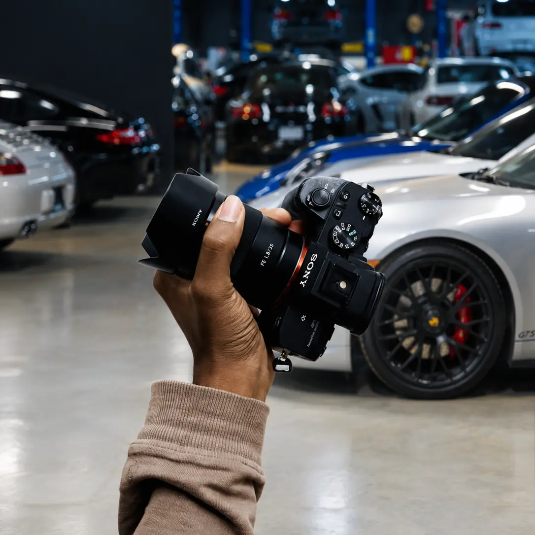 Hand holding a Sony camera in the M Sport Motorwerx garage, Porsches behind